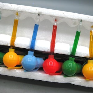 Könnte beinhalten: Eine Lichterkette mit Vintage-Weihnachtslichtern mit bunten Glühbirnen und Glasröhren, die mit Flüssigkeit gefüllt sind. Die Glühbirnen sind in verschiedenen Farben gehalten, darunter Grün, Gelb, Blau und Rot, und sind in einem weißen Schaumstoffhalter angeordnet.
