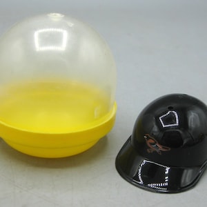 Op de afbeelding: Een doorzichtige plastic capsule met een gele basis ligt naast een zwarte baseballpet. De pet heeft een gebogen klep en een klein vogel-logo. De capsule is transparant.