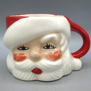 May include: A ceramic mug shaped like Santa Claus's face. The mug features a red handle and rim, a white beard and hat, and a rosy-cheeked face. The mug is a festive holiday item.