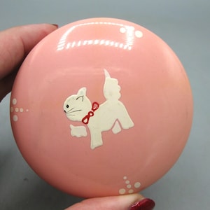 May include: A round, pink decorative object featuring a white cat illustration with a red bow. The cat is centered, with white dots on the side. The object is held by a hand.