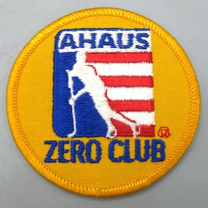 May include: A round, yellow patch with a blue, white, and red design. The patch features the text "AHAUS" above a hockey player silhouette and the text "ZERO CLUB" in blue. The patch has a stitched border.