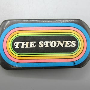 Puede incluir: Un objeto metálico rectangular vintage con fondo negro y el texto "THE STONES" en blanco. El texto está rodeado por una serie de formas ovaladas concéntricas de colores azul, amarillo, naranja, rosa y verde.