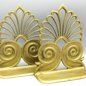 May include: A pair of antique gold-coloured bookends. Each bookend has a stylized fan-shaped top and a large, swirling scroll design. The bookends are made of metal and have a rectangular base.