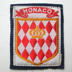 Op de afbeelding: Geborduurde patch met het woord "MONACO" in rood op een witte banner. De patch heeft een schildontwerp met een rood-wit ruitpatroon, een gouden kroon en een gouden rand, alles tegen een marineblauwe achtergrond.