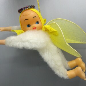 May include: A hanging ornament featuring a doll with a peach-toned face, blue eyes, and red lips, wearing a yellow hood with a multicolored tinsel halo. The doll has a white fuzzy body, yellow mesh wings, and peach-toned arms and feet.