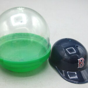 Op de afbeelding: Een miniatuur marineblauwe baseballpet met een rood "B"-logo, naast een doorzichtige plastic capsule met een groene basis. De pet heeft een glanzende afwerking en is een verzamelobject.