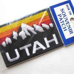 Peut inclure: Un écusson souvenir thermocollant avec le mot "UTAH" en broderie blanche. Le patch a un fond noir avec un motif de chaîne de montagnes en blanc, jaune, orange et rouge. Le patch est dans un emballage plastique transparent.