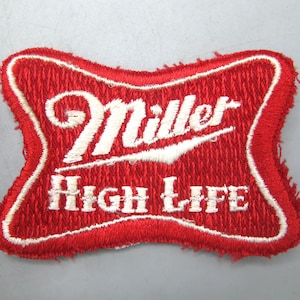 May include: A red embroidered patch with the words "Miller High Life" in white. The patch has a distinctive shape and a white border. The text is in a stylized font, and the patch has a textured look.