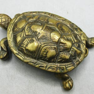 Può includere: Una statuetta di tartaruga in ottone caratterizzata da un guscio dettagliato e strutturato con un motivo geometrico, una piccola testa con segni incrociati e quattro zampe estese. La superficie metallica ha una finitura dorata lucida.