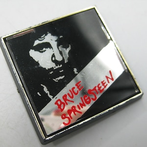 May include: Square pin with a black background and a silver diagonal stripe. The pin features a white silhouette of a person's face and the words "BRUCE SPRINGSTEEN" in red.