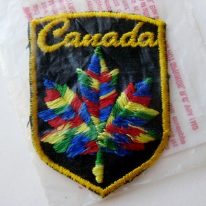 May include: Embroidered patch featuring the word "Canada" in yellow script above a colorful maple leaf. The leaf is stitched with red, blue, green, and yellow threads. The patch has a black background and a yellow border.