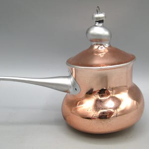 Op de afbeelding: Een glazen kerstboomornament in de vorm van een koperkleurige steelpan met een zilveren handvat en een zilveren dekselknop. Het ornament heeft een reflecterende metallic afwerking en een klein zilveren oogje om in de boom te hangen.