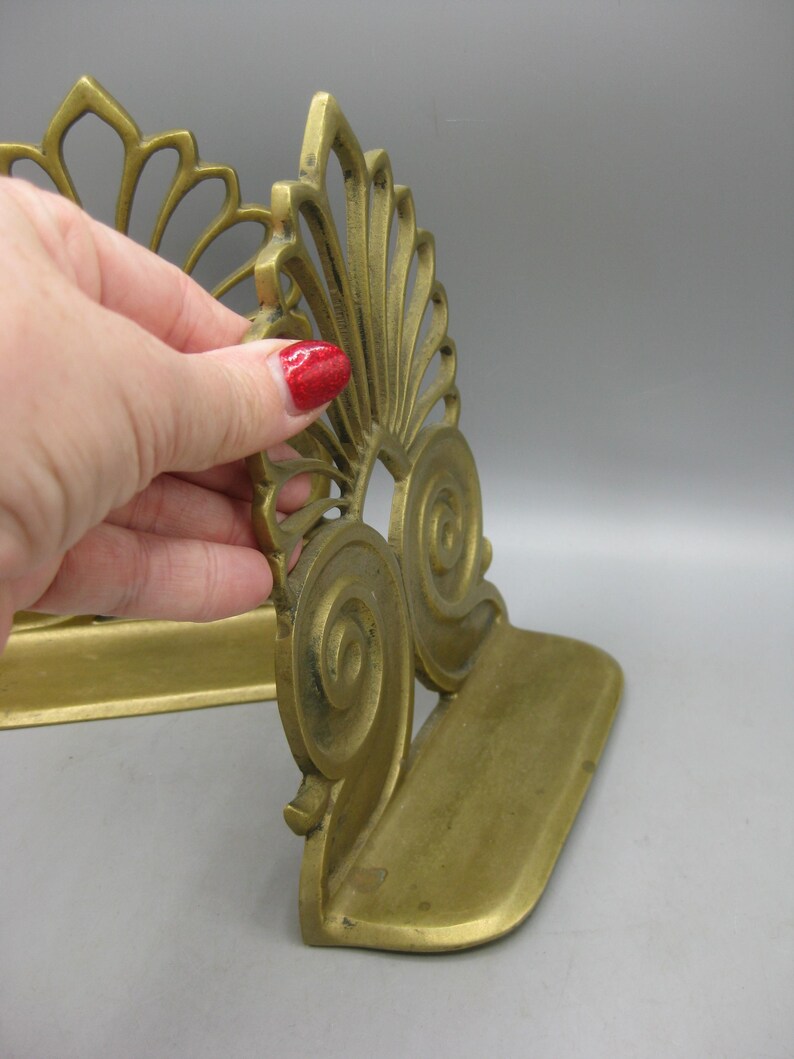 May include: A pair of antique brass bookends. Each bookend features an ornate, fan-shaped design with scroll details. The bookends are a warm, golden colour and appear to be made of solid brass. The base is approximately 10 cm long.
