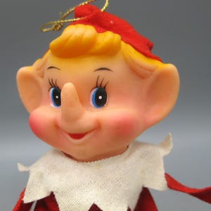 Peut inclure: Une poupée elfe vintage avec une expression joyeuse. La poupée a un teint clair, des joues roses et des yeux bleus. Elle porte un chapeau et une tenue rouges avec un col blanc. Un halo en fil d'or est posé sur le chapeau.