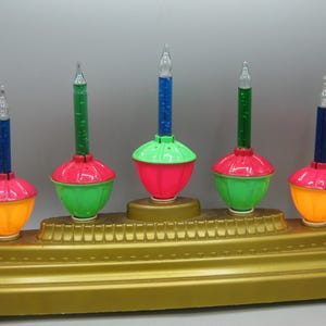 Op de afbeelding: Een vintage kerstverlichting display met vijf gekleurde bubbellampen. Elke lamp heeft een basis in oranje, rood en groen, met een heldere, puntige top. De lampen zijn gemonteerd op een goudkleurige basis.