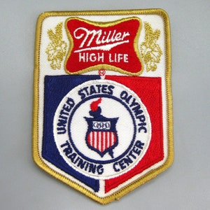 May include: Embroidered patch with the Miller High Life logo above the United States Olympic Training Centre emblem. The patch has a white background with red, blue, and gold accents. The text "Miller High Life" is in red script.