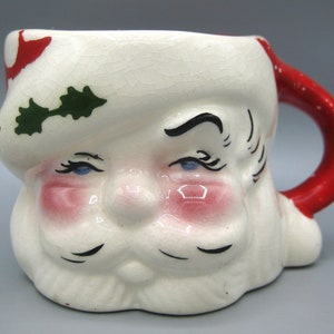 May include: A ceramic Santa Claus face mug with a red handle. The mug is painted with a white base, red accents, and pink cheeks. The Santa face has blue eyes, a white beard, and a red hat with green holly.