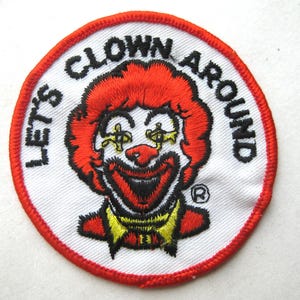 Puede incluir: Un parche bordado circular que presenta un payaso con cabello rojo brillante, cuello amarillo y una amplia sonrisa. El texto "LET'S CLOWN AROUND" está bordado en letras negras a lo largo del borde superior del fondo blanco, con un borde rojo.
