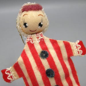 May include: A vintage doll ornament with a fabric face and a red and white striped outfit. The doll has a small red hat and a white collar with lace trim. The outfit has two dark blue pom-pom buttons. The doll's face has painted eyes and a small smile.