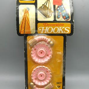May include: Vintage package of two pink decorative hooks with gold accents. The package features the brand name "nevco" and the text "Decorative Hooks". The package also shows images of items hanging on hooks.