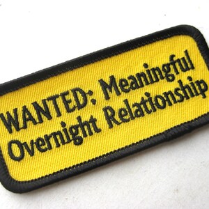 Puede incluir: Un parche rectangular bordado con un fondo amarillo brillante y un borde negro grueso. El texto "WANTED: Meaningful Overnight Relationship" está bordado con hilo negro en el centro del parche en una fuente sans-serif clara.