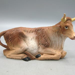 May include: A brown and white figurine of a cow in a resting position. The cow has yellow horns, black hooves, and a red mouth. The figurine is likely part of a nativity scene.