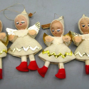 Peut inclure: Quatre décorations d'anges vintage. Chaque ange porte une robe en feutre crème avec des bordures dorées et argentées, des ailes dorées et des bottes en feutre rouge. Les anges ont les yeux fermés et portent des capuches blanches.