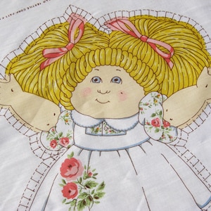 May include: A fabric panel featuring a doll with blonde hair styled in pigtails, each adorned with a pink bow. The doll has blue eyes, rosy cheeks, and is wearing a white dress with a floral pattern. The edges are outlined with a dashed stitch.