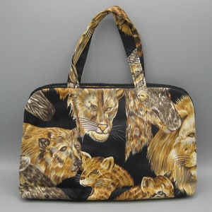 May include: A black handbag featuring a repeating pattern of lions and lionesses in brown and tan hues. The bag has a rectangular shape, a zip closure, two handles, and a black cord trim.