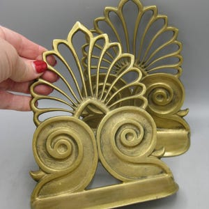 May include: A pair of antique brass bookends, featuring a fan-shaped top and a large scroll design at the base. The bookends have a warm gold hue and a vintage appearance. They are set against a grey backdrop.