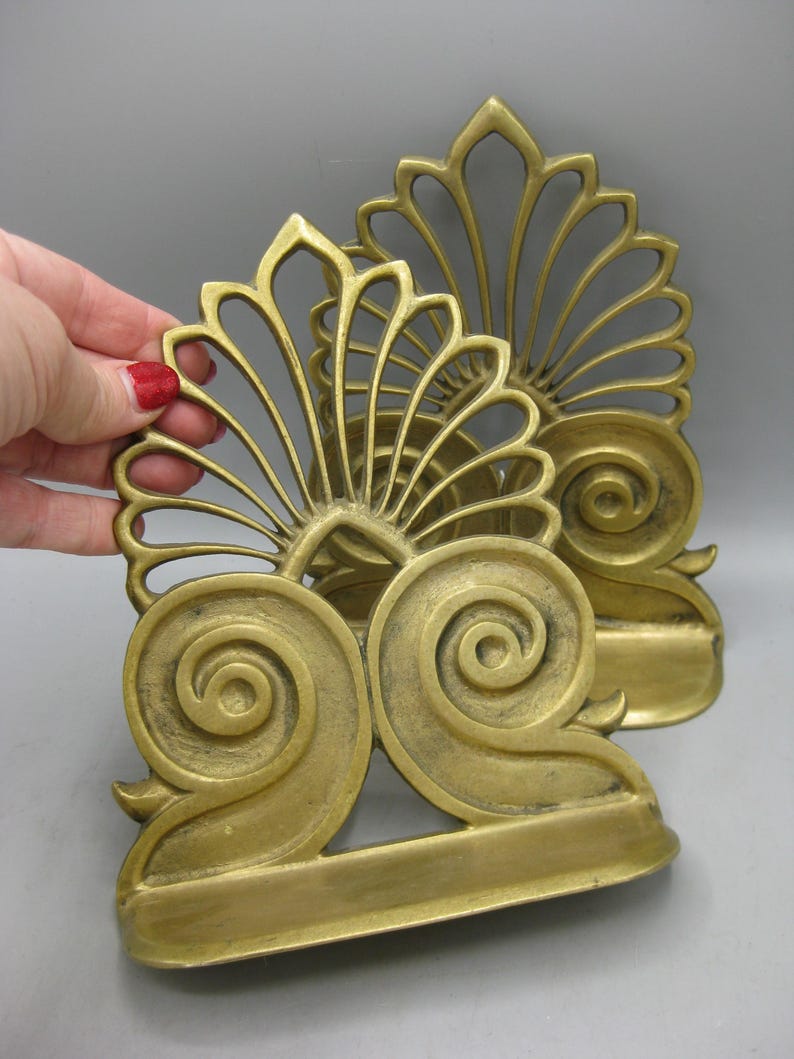 May include: A pair of antique brass bookends, featuring a fan-shaped top and a large scroll design at the base. The bookends have a warm gold hue and a vintage appearance. They are set against a grey backdrop.