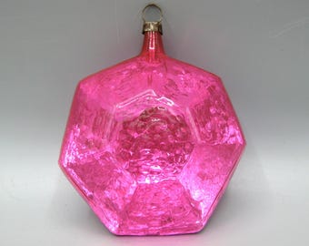 Op de afbeelding: Een felroze, zeshoekig glazen ornament met een getextureerd, kuiltjesoppervlak. Het ornament heeft een zilverkleurige metalen dop aan de bovenkant met een kleine ronde lus om op te hangen, die het licht reflecteert over het gefacetteerde glazen lichaam.