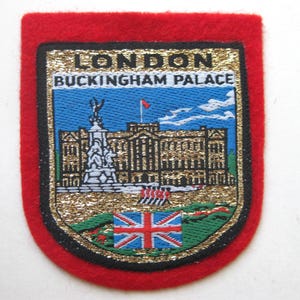 May include: A red, embroidered patch featuring a detailed image of Buckingham Palace in London, with the text "LONDON BUCKINGHAM PALACE". The patch includes a Union Jack flag and a gold glitter border.
