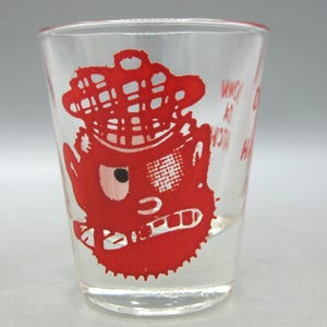 Peut inclure: Verre à shot transparent avec un dessin de personnage de dessin animé rouge. Le personnage porte un chapeau, un seul œil et un sourire édenté. Le verre a du texte sur le côté. Un objet de collection vintage.