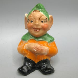 May include: A ceramic figurine of a smiling elf with a green hat and collar, an orange shirt, and black boots. The elf has rosy cheeks and is holding its hands together.
