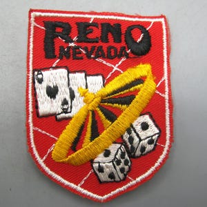 May include: Embroidered patch with a red background and white border. The patch features the text "RENO NEVADA" in black, along with images of playing cards, dice, and a roulette wheel. The roulette wheel is yellow and black.