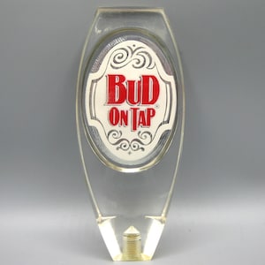 Könnte beinhalten: Transparenter Bud On Tap Zapfhahn. Der Griff hat eine ovale Fläche mit den Worten "Bud On Tap" in roter Schrift auf weißem Hintergrund. Der Griff hat ein dekoratives Design um den Text herum.