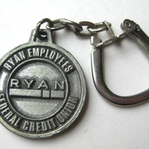 May include: A silver-colored keychain featuring a circular pendant with the text "RYAN EMPLOYEES FEDERAL CREDIT UNION." The pendant is connected to a chain and a metal clasp. The word "RYAN" is prominently displayed.