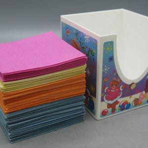 May include: A stack of colorful square sticky notes in pink, yellow, orange, and blue, next to a white plastic holder with a whimsical design featuring a bear, reindeer, and other festive elements.