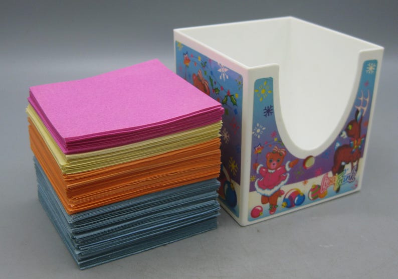 May include: A stack of colorful square sticky notes in pink, yellow, orange, and blue, next to a white plastic holder with a whimsical design featuring a bear, reindeer, and other festive elements.