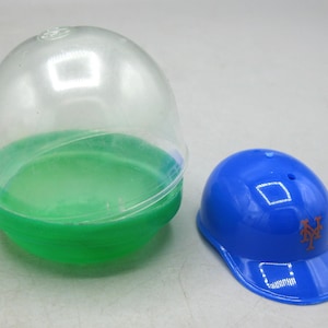 Op de afbeelding: Een blauwe baseballpet met het New York Mets-logo naast een doorzichtige plastic capsule met een groene basis. De pet is glanzend en heeft een gebogen klep. De capsule is open en toont een groen interieur.