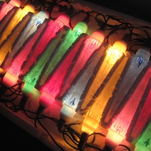 May include: A string of vintage Christmas lights with colorful bulbs. The bulbs are shaped like icicles and emit a warm, inviting glow. The lights are arranged in a box, creating a festive display. The colors include red, green, yellow, and white.