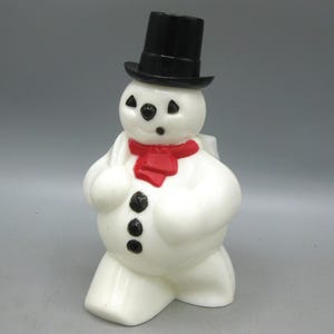 Può includere: Una statuetta di pupazzo di neve in plastica bianca che indossa un cilindro nero e una sciarpa rossa. Il pupazzo ha occhi, naso e una piccola bocca dipinti di nero, con tre bottoni neri sul davanti del busto. È raffigurato mentre cammina.