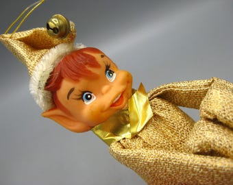 May include: A vintage Christmas elf ornament with a smiling face, red hair, and blue eyes. The elf wears a gold hat with a bell and a gold outfit with a gold ribbon. A festive holiday decoration.