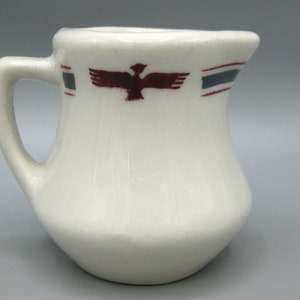 May include: Cream-colored ceramic creamer with a handle and a flared spout. The creamer features a maroon bird design and a decorative band of maroon and gray stripes near the rim. A small, vintage-style pitcher.