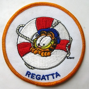 May include: A circular embroidered patch with an orange border featuring a white and red life preserver. Inside the life preserver is an orange tabby cat wearing a blue hat. A white rope wraps around the life preserver. The word REGATTA is in blue.