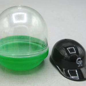 Op de afbeelding: Een zwarte honkbalhelm met een wit "CR" logo naast een doorzichtige plastic capsule met een groene basis. De helm is glanzend en heeft een kleine klep. De capsule is leeg.