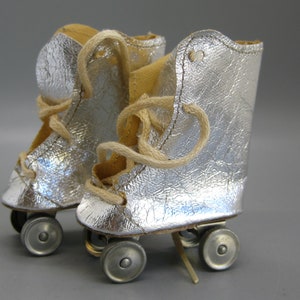 May include: A pair of miniature silver roller skates with beige laces. The skates have a textured, metallic appearance and are set against a neutral background. The wheels are small and silver, and the laces are tied.