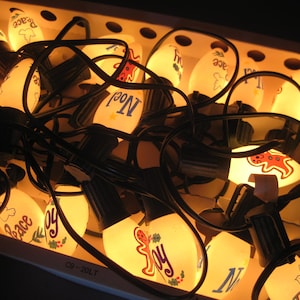 May include: A string of vintage Christmas lights with white bulbs. Each bulb is decorated with holiday-themed images and words like "Joy", "Peace", and "Noel". The bulbs are connected by black wires and are in a cardboard box.