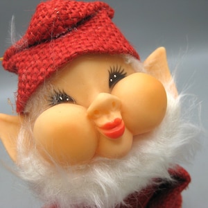 May include: A close-up of a vintage gnome figurine. The gnome has a red burlap hat, a white fluffy beard, and a smiling face with rosy cheeks and red lips. The figurine is a whimsical decorative item.